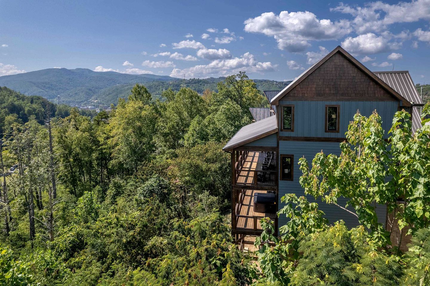 A two-story house with a balcony, surrounded by dense forest and mountains in the background.