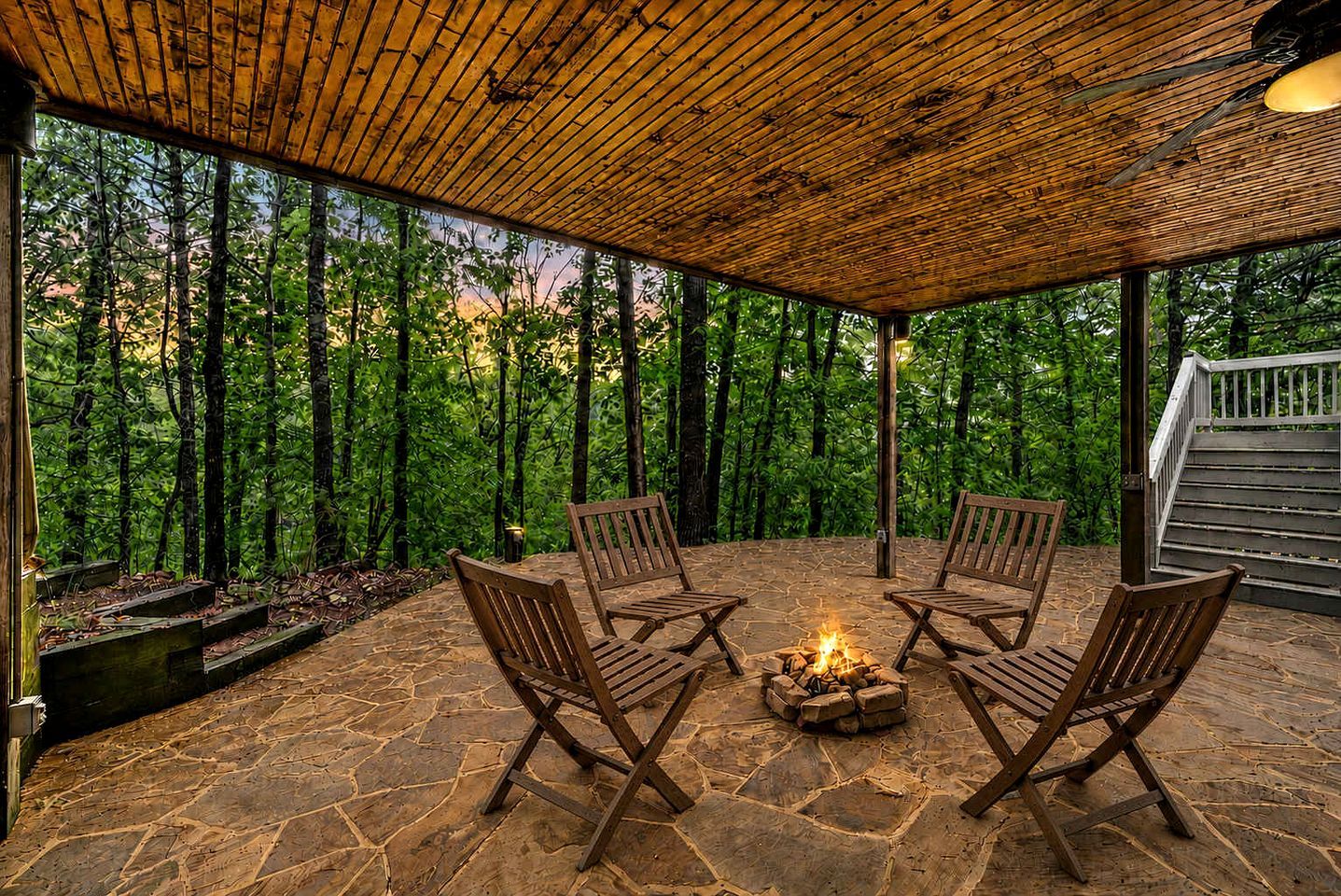 A covered outdoor seating area with stone flooring, wooden chairs, and a fire pit.
