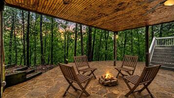 A covered outdoor seating area with stone flooring, wooden chairs, and a fire pit.