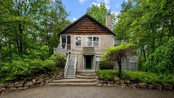 A two-story wooden house with a stone staircase leading to the front door, surrounded by lush greenery.