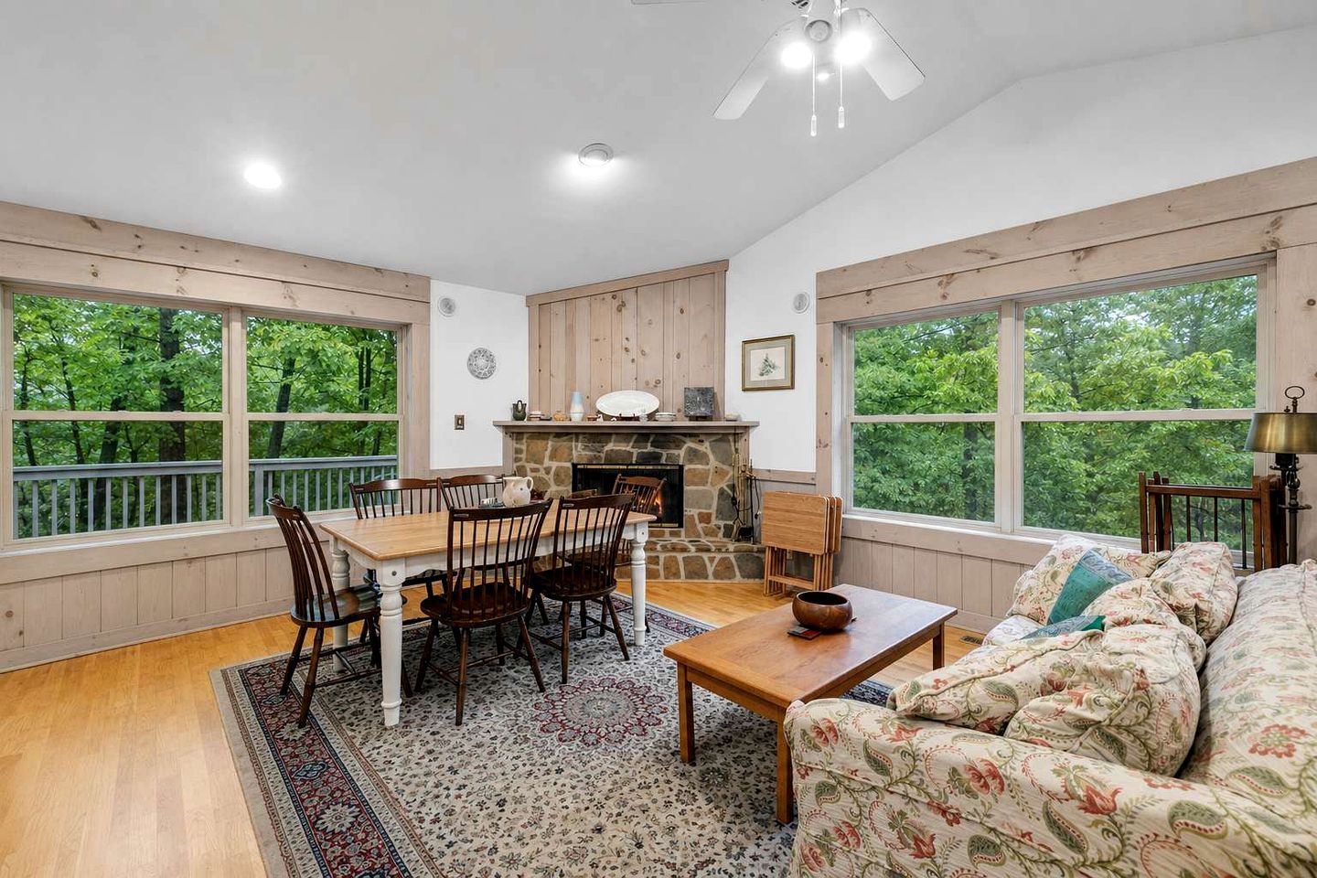 A cozy living room with a fireplace, wooden furniture, and large windows overlooking greenery.