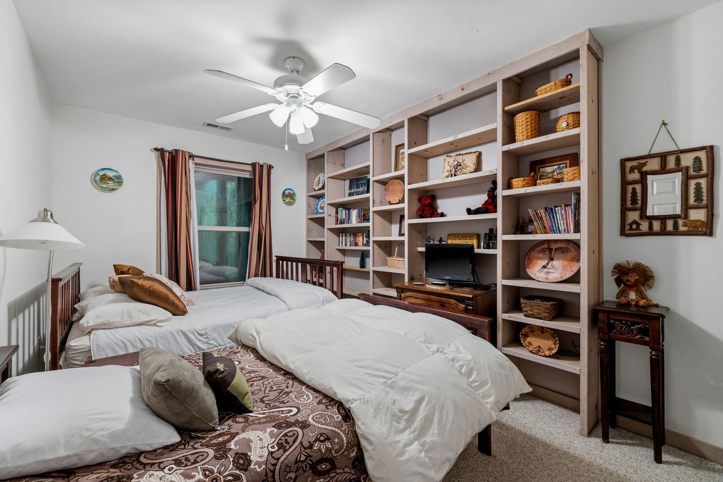 A bedroom with two beds, a ceiling fan, a bookshelf, and a window with curtains.