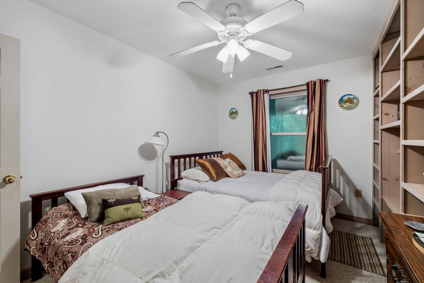 Une chambre avec deux lits, un ventilateur de plafond, une fenêtre avec des rideaux et une bibliothèque.