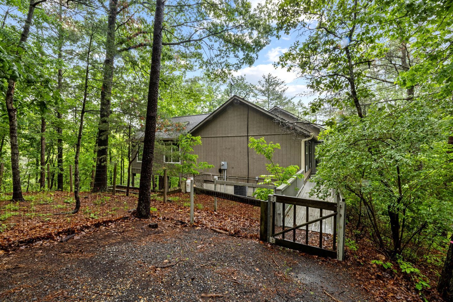 A rustic cabin surrounded by a forest, with a gated entrance and a gravel driveway.