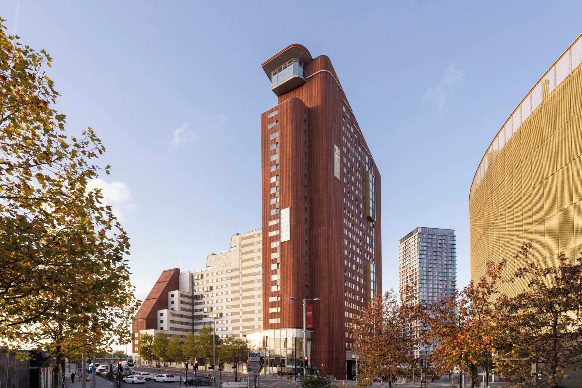 A tall red-brick building with a unique top section, surrounded by other modern buildings and trees with autumn leaves.