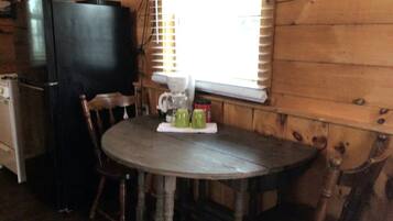 Um interior de madeira com uma mesa redonda, cadeiras, um frigorífico preto e uma janela com persianas.