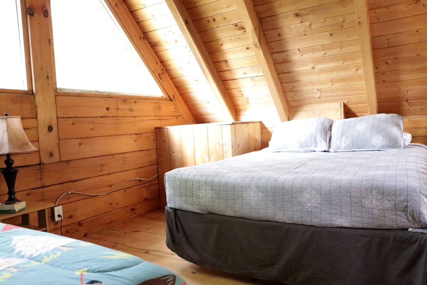 Une chambre à coucher dans une cabane en bois, avec un plafond en pente, un lit recouvert d’une courtepointe matelassée, une lampe de chevet et une fenêtre.
