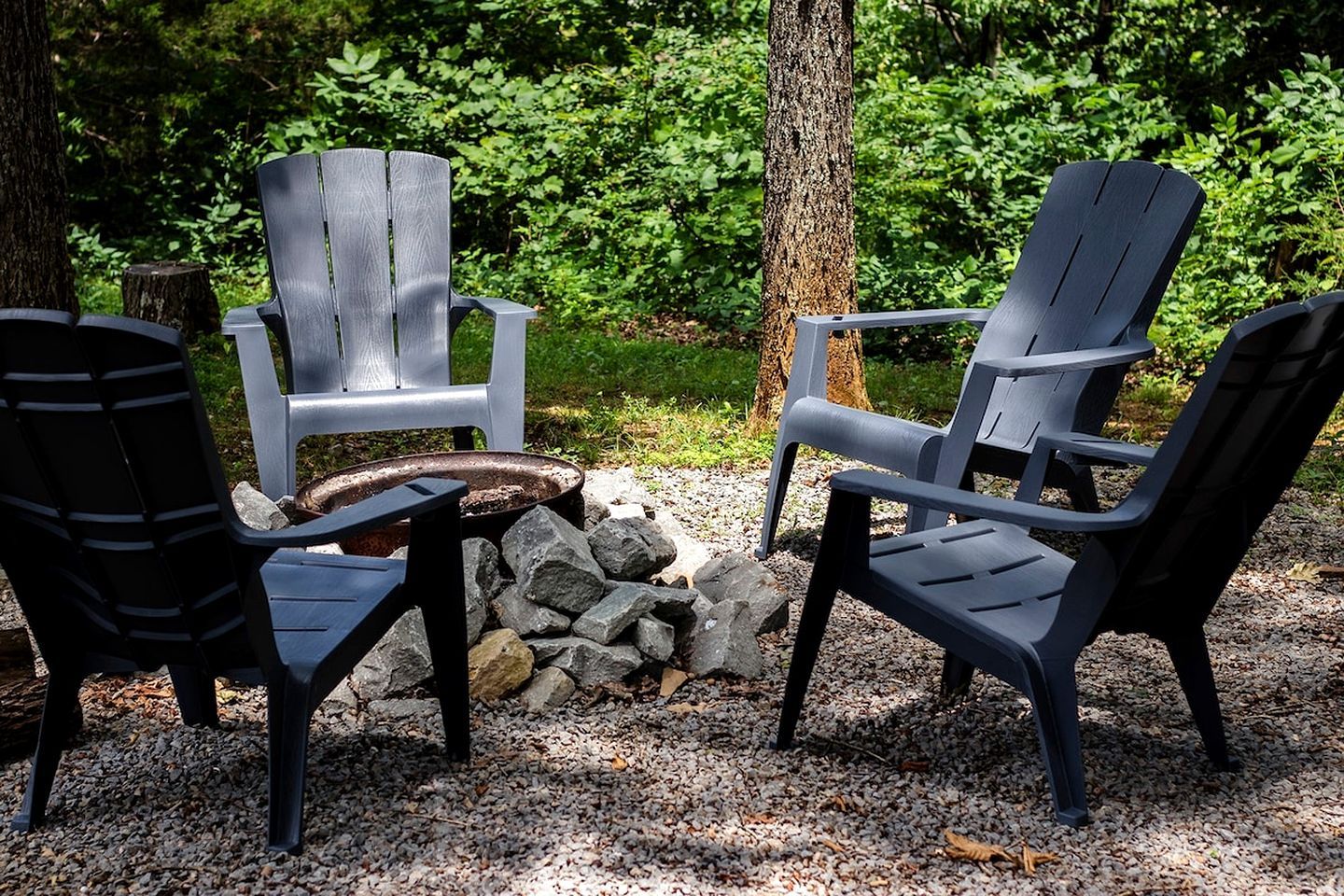 Two Adirondack chairs with a fire pit between them, surrounded by rocks and greenery.