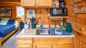 A cozy cabin interior with a bed, a kitchen area featuring wooden cabinets, a microwave, and a coffee station.