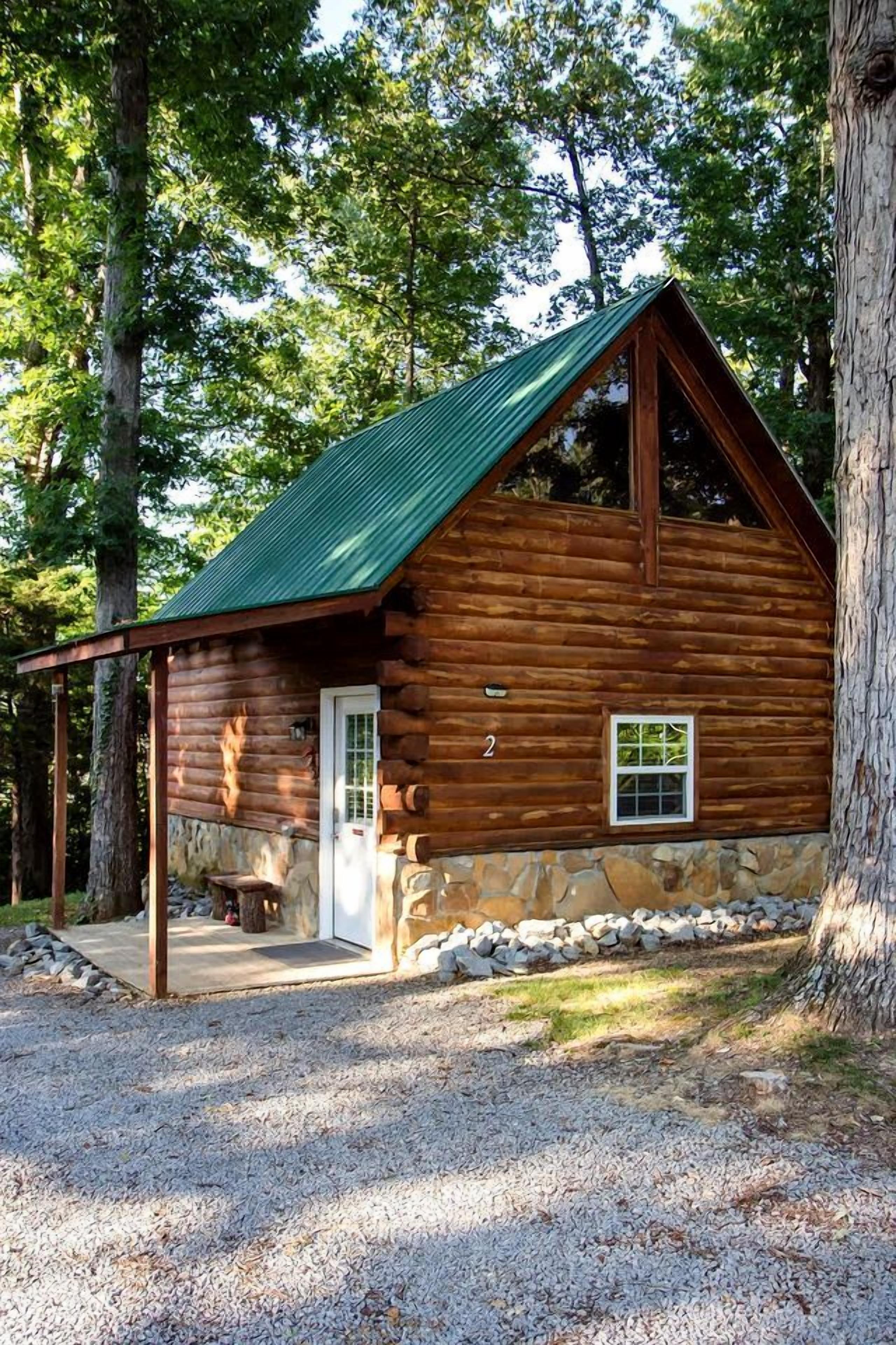 A log cabin with a stone foundation, a green metal roof, and a gravel driveway.