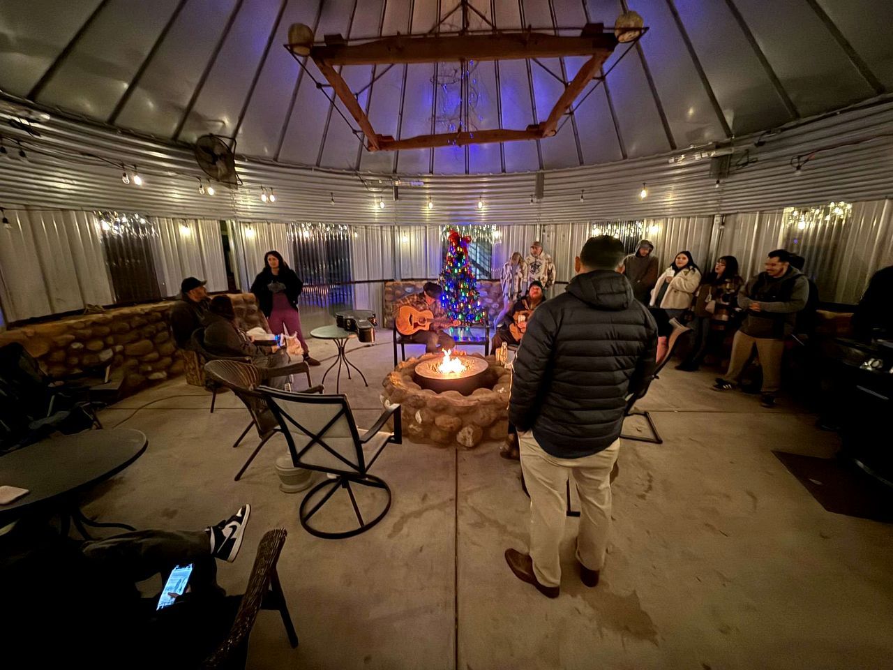 An indoor venue with a fire pit, tables, chairs, and a Christmas tree.