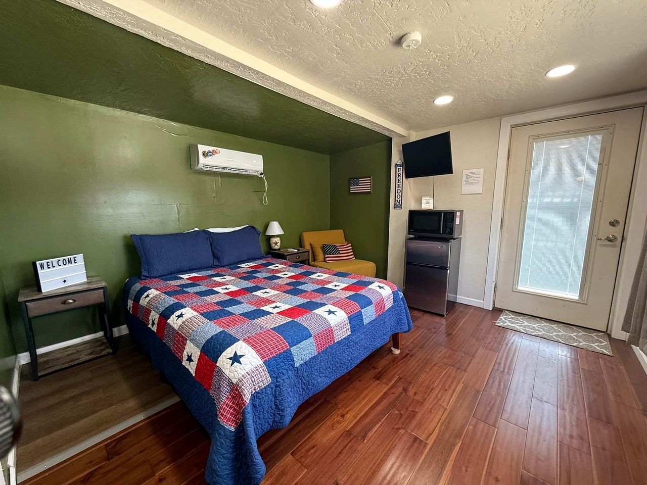 A bedroom with a bed, a nightstand, a lamp, a small table, a flag, a refrigerator, and a door with a window.