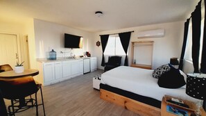 A compact hotel room with a kitchenette, a bed with patterned pillows, a dining area with a table and chairs, and a large window with curtains.