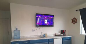 A kitchen with a large water dispenser, a coffee maker, and a wall-mounted television displaying a cityscape.