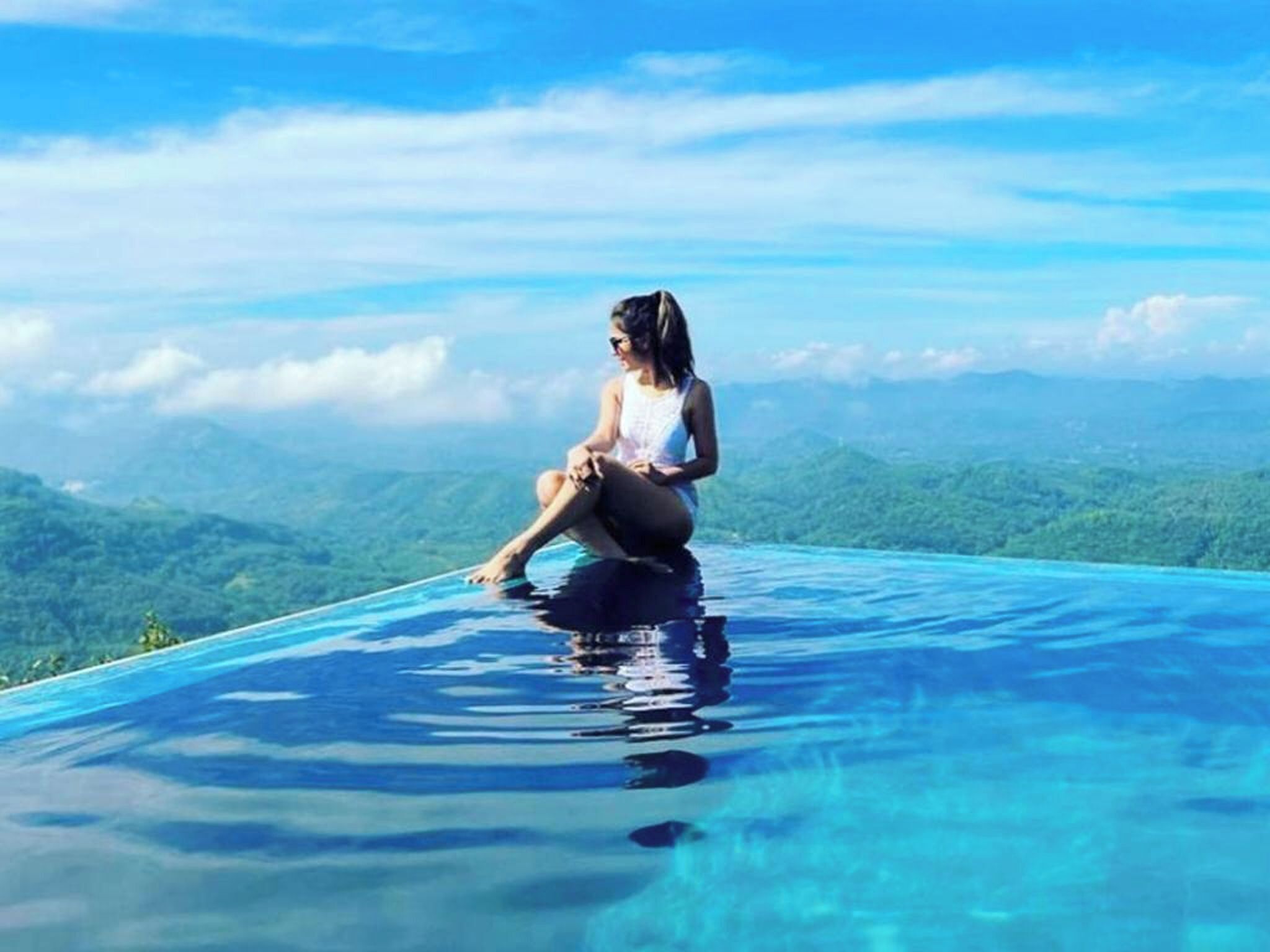 Una persona sentada al borde de una piscina infinita azul cristalina, con un paisaje montañoso de fondo.