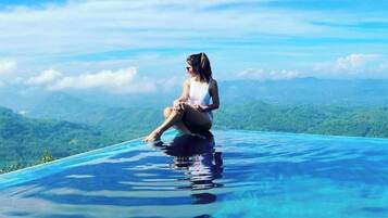 Una persona sentada al borde de una piscina infinita azul cristalina, con un paisaje montañoso de fondo.