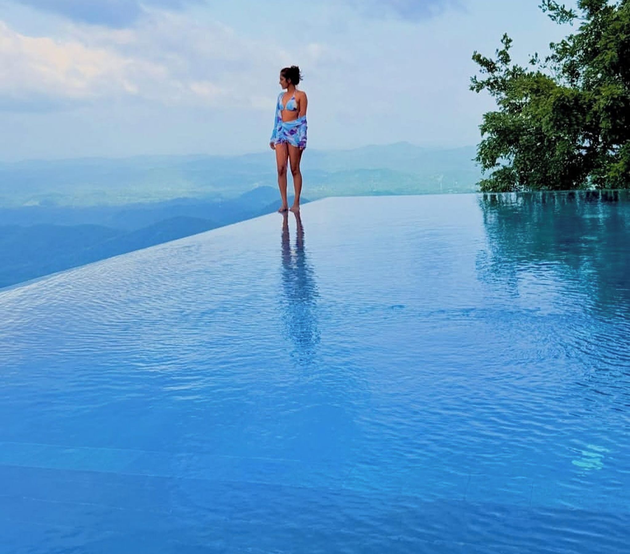 Una persona está de pie en una piscina infinita azul cristalina con vistas a montañas lejanas.