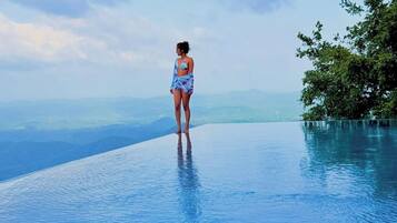 Una persona está de pie en una piscina infinita azul cristalina con vistas a montañas lejanas.
