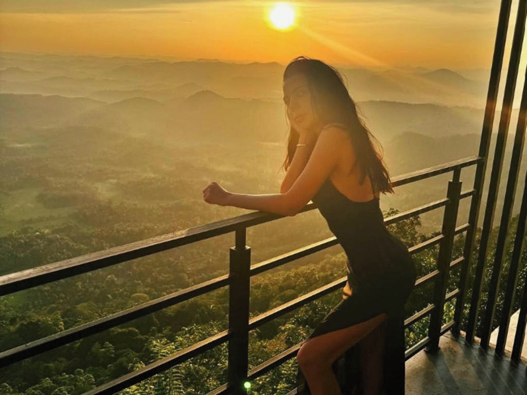 Una persona apoyada en la barandilla de un balcón, con vistas a un paisaje montañoso al atardecer.