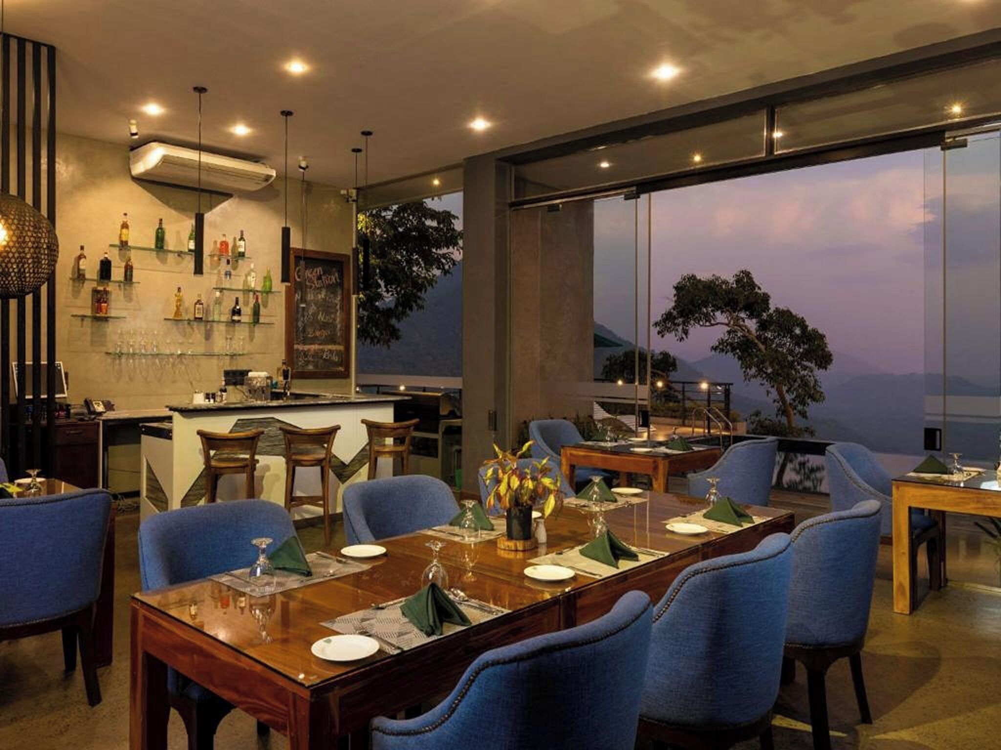 Un comedor moderno con una mesa larga preparada para una comida, rodeada de sillas azules, y vistas a las montañas a través de amplios ventanales.
