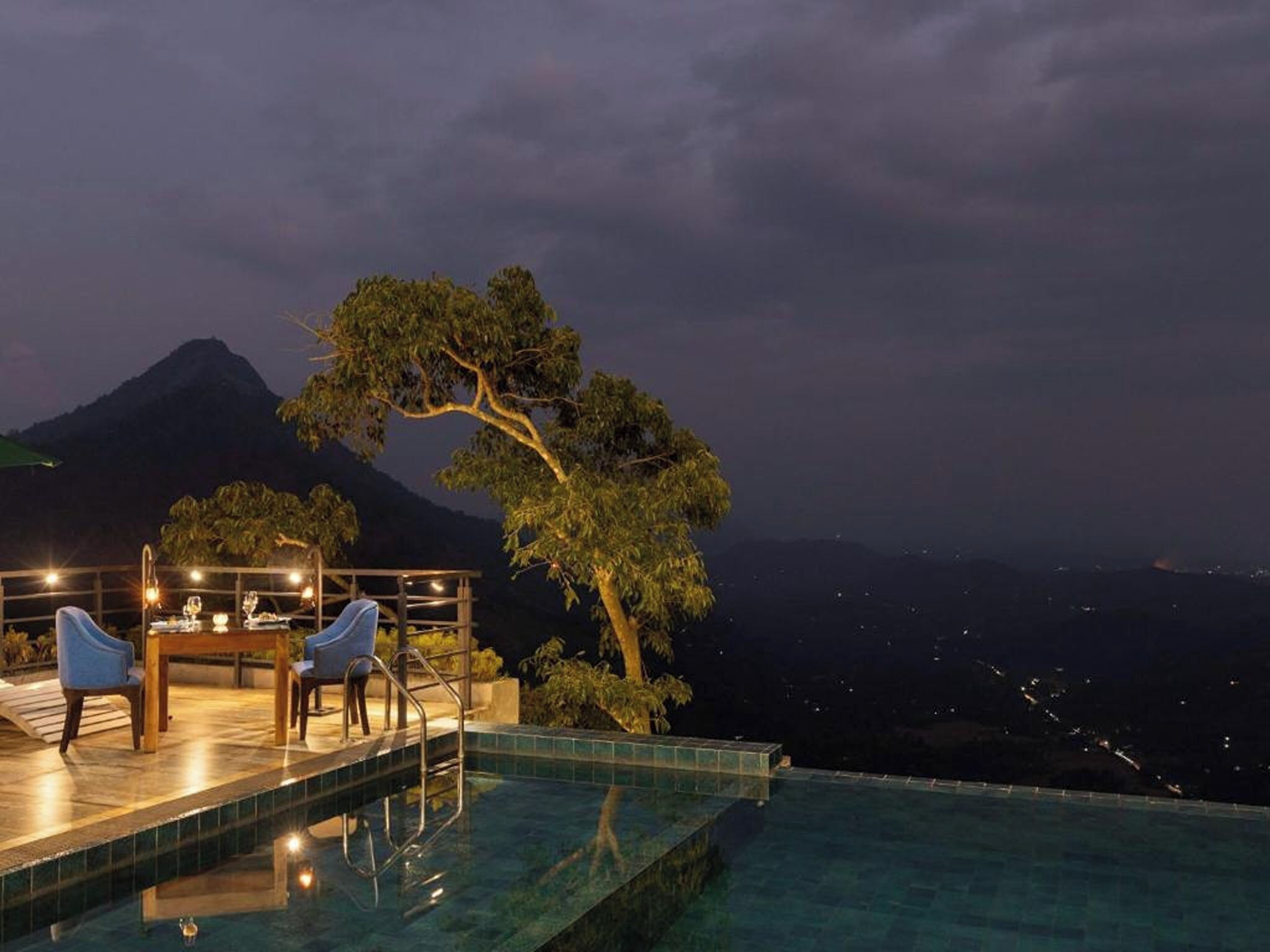 An infinity pool with a dining area and chairs, overlooking a mountainous landscape at night.