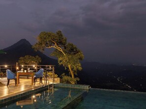 Una piscina infinita con zona de comedor y sillas, con vista a un paisaje montañoso de noche.