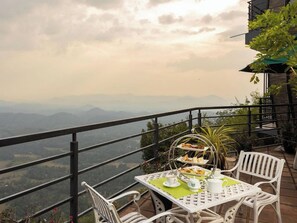A table set with a tea set and a tiered dessert stand on a balcony overlooking a mountainous landscape.