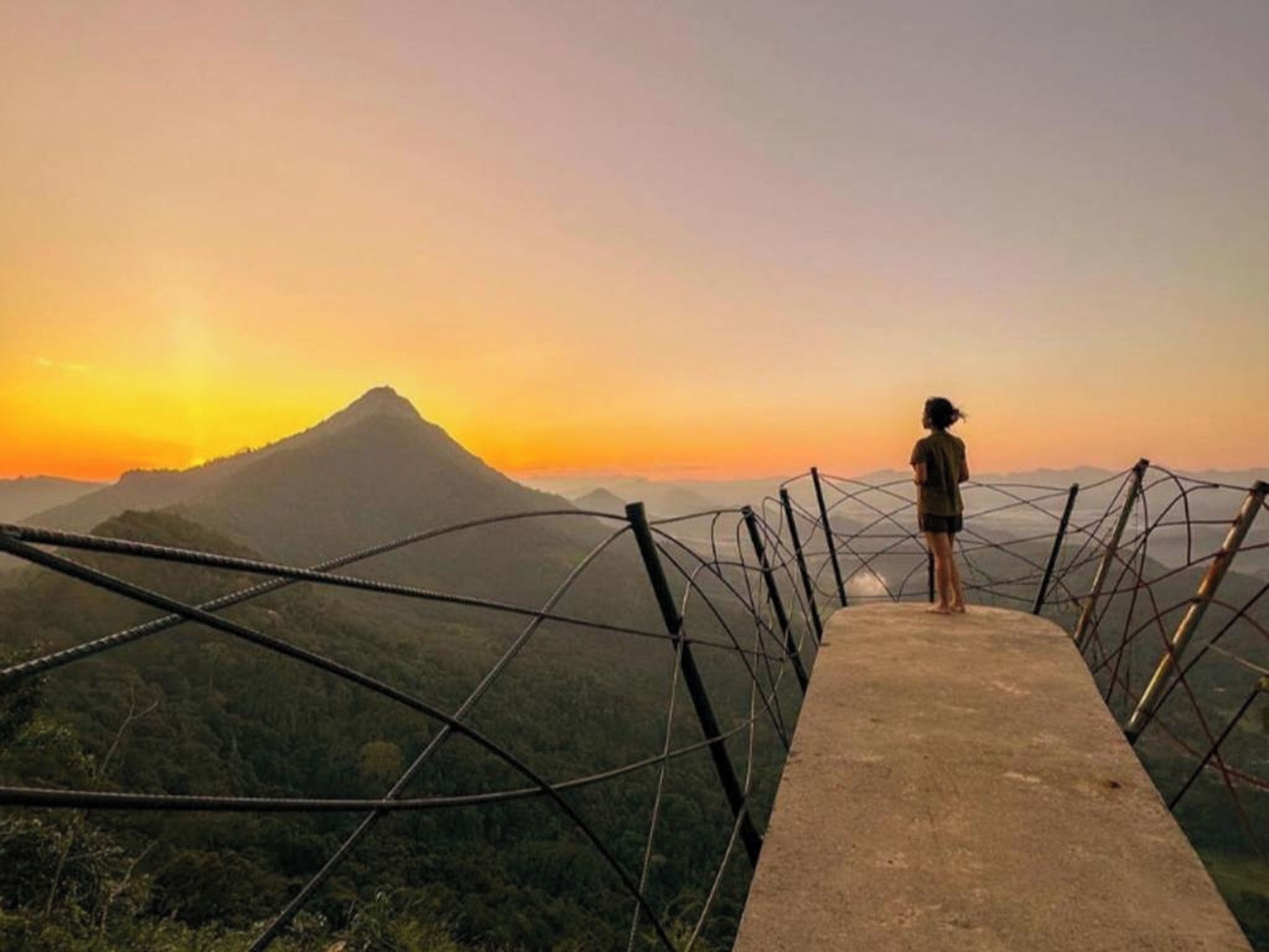 A person standing on a concrete platform with a metal fence, overlooking a mountainous landscape during sunset.