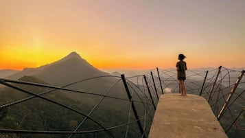 A person standing on a concrete platform with a metal fence, overlooking a mountainous landscape during sunset.