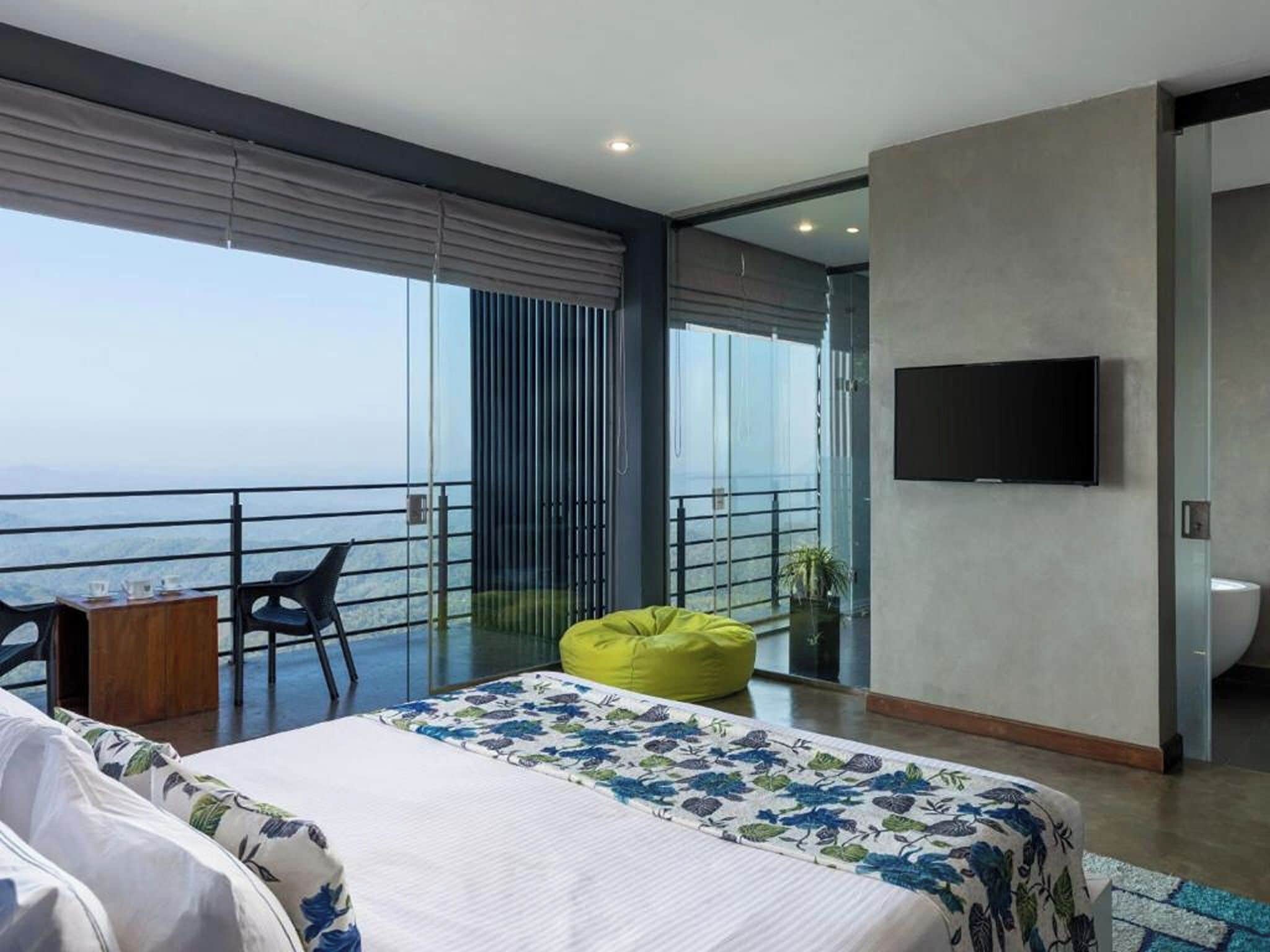 Quarto de hotel moderno com uma cama grande, varanda com vista, televisão de tela plana e uma pequena mesa com cadeiras.