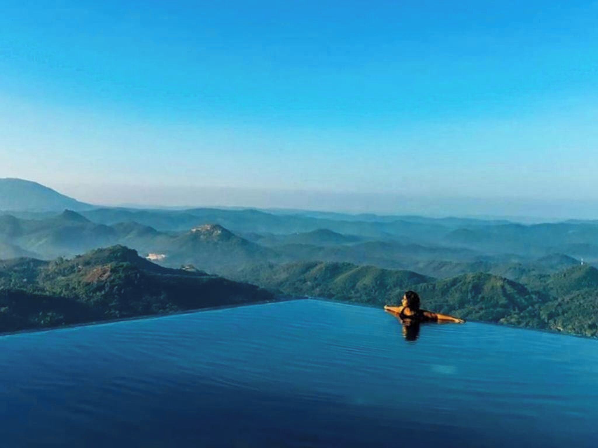 Uma pessoa relaxando em uma piscina infinita azul cristalina, com uma paisagem montanhosa ao fundo.