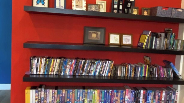 A bookshelf with various items including books, DVDs, and framed photos against a red wall. - Urban Living in Kampung Setting (Kuching)
