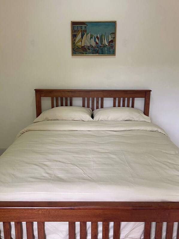 A bed with a wooden headboard, a painting of sailboats on the wall, and a plain white wall. - Urban Living in Kampung Setting (Kuching)
