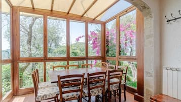 A dining area with a large table and chairs, large windows with a view of trees and flowers, and a stone wall.