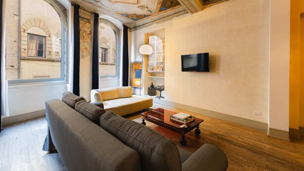 Un salon comprenant un canapé, une table basse avec des livres, un téléviseur fixé au mur et une grande fenêtre donnant sur un immeuble.