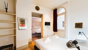 Une chambre à coucher comprenant un lit, un bureau, une chaise, une télévision, une lampe et une salle de bain visible à travers une porte ouverte.