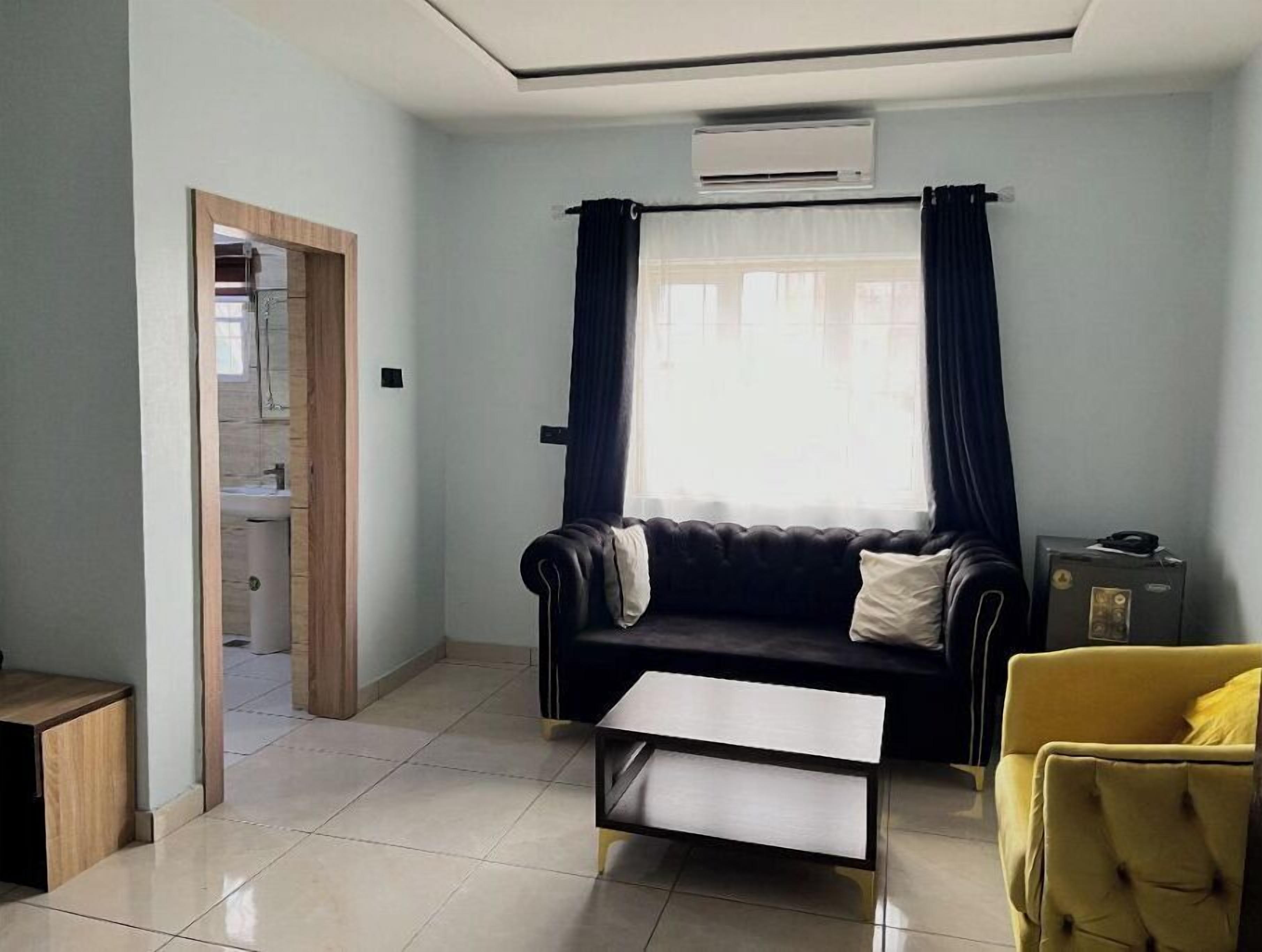 A living room with a sofa, a coffee table, a yellow armchair, a window with curtains, and an air conditioning unit.