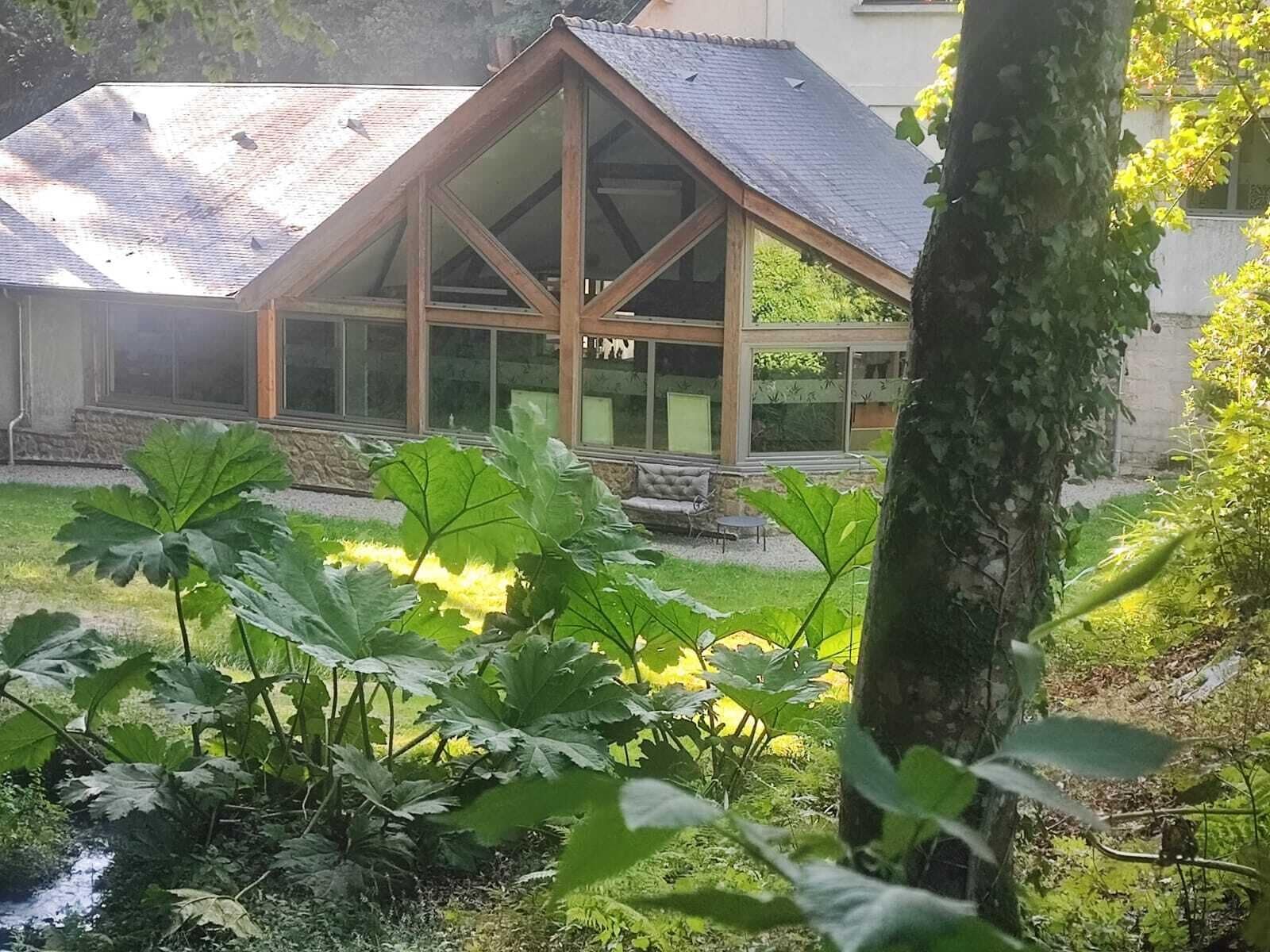 A building with a sloped roof and large windows, surrounded by lush greenery and a small pond.