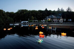 Ein ruhiger See mit schwebenden Laternen, einer Anlegestelle und einem Gebäude, vor dessen Ufer sich Menschen befinden.