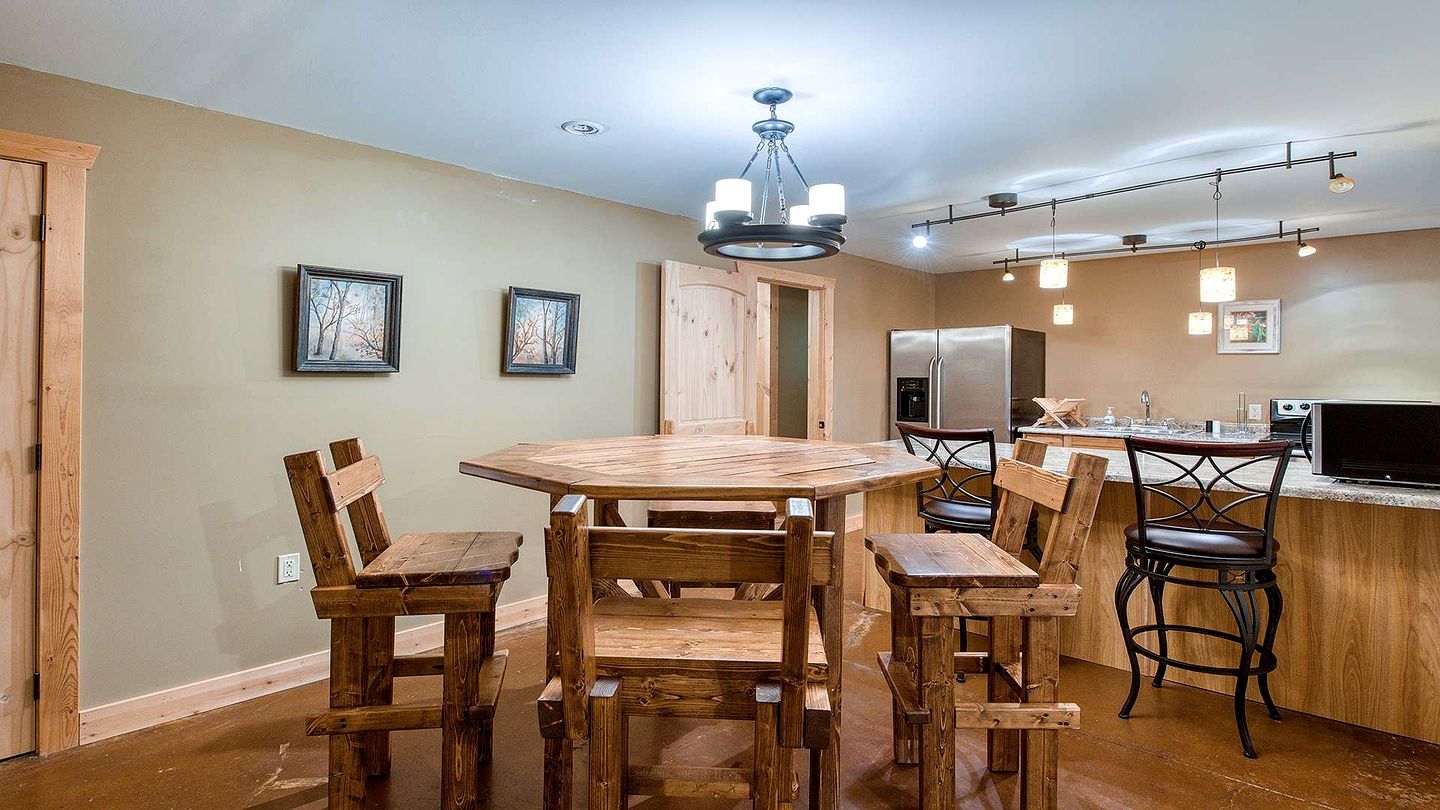 A dining area with a wooden table and chairs, a chandelier, and a kitchen with a refrigerator and microwave.