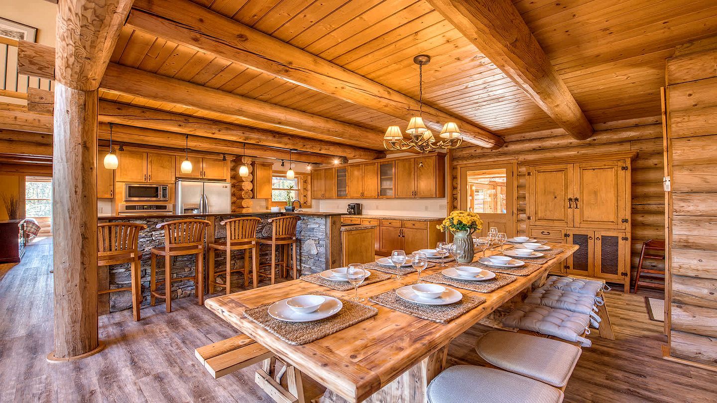 Interior de una cabaña de madera con una larga mesa de comedor para seis personas, zona de cocina con electrodomésticos y detalles de piedra.