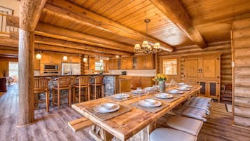 Interior de una cabaña de madera con una larga mesa de comedor para seis personas, zona de cocina con electrodomésticos y detalles de piedra.