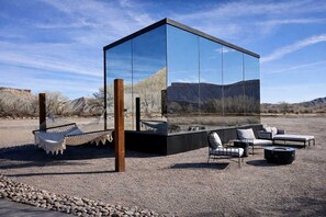 A modern glass structure with a hammock, lounge chairs, and a fire pit.