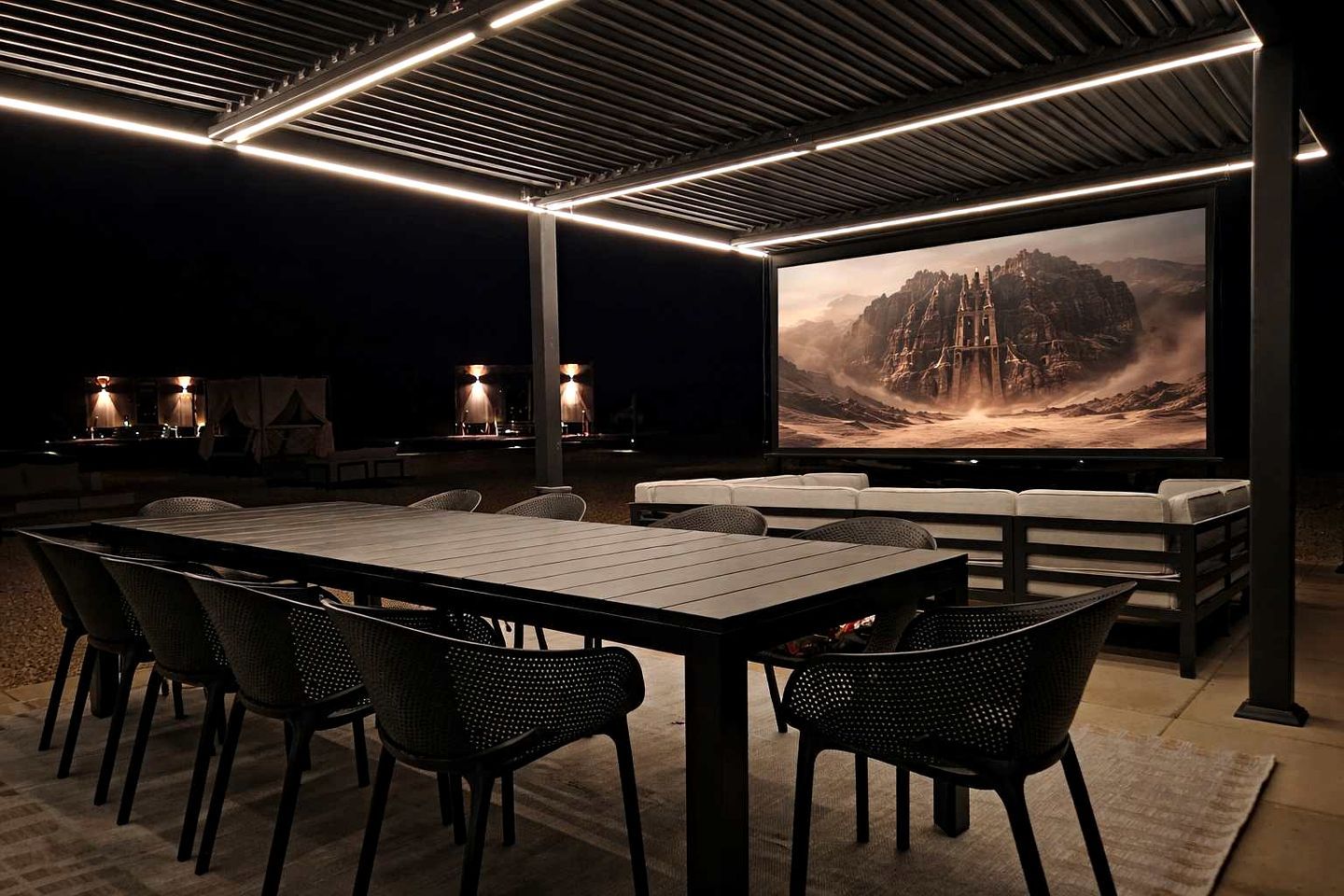 An outdoor dining area with a large table and chairs, a flat-screen TV displaying a mountainous landscape, and ambient lighting.