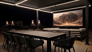 An outdoor dining area with a large table and chairs, a flat-screen TV displaying a mountainous landscape, and ambient lighting.