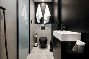 A modern bathroom with a glass shower enclosure, a toilet, a sink with a towel rack, and a trash bin.