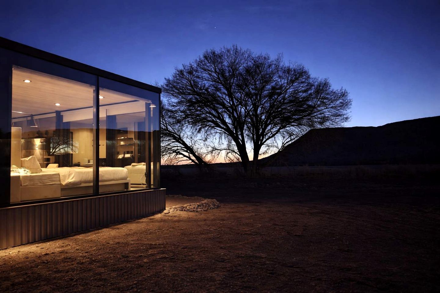 A modern glass structure with a bed and seating area, surrounded by a natural landscape.