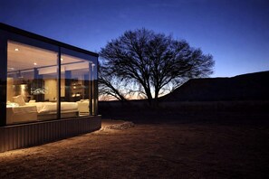 A modern glass structure with a bed and seating area, surrounded by a natural landscape.