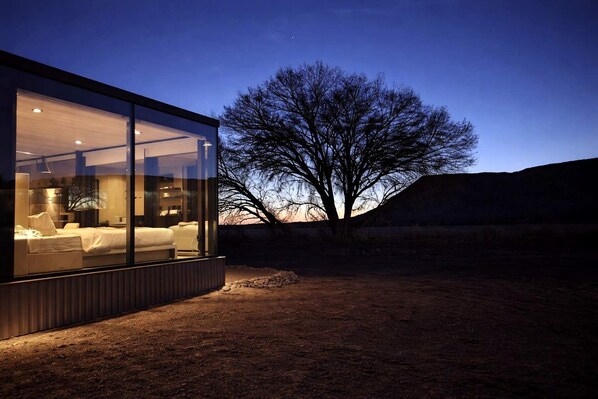 A modern glass structure with a bed and seating area, surrounded by a natural landscape.