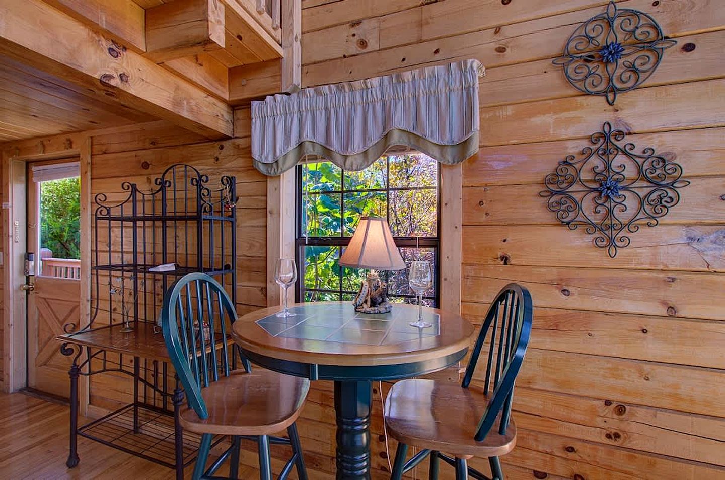 A wooden interior with a round table, two chairs, a window with a view, and decorative metal wall art.